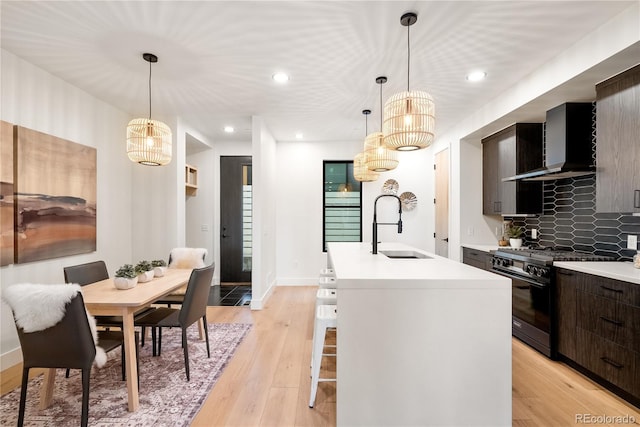 kitchen with sink, hanging light fixtures, black gas range oven, an island with sink, and wall chimney range hood