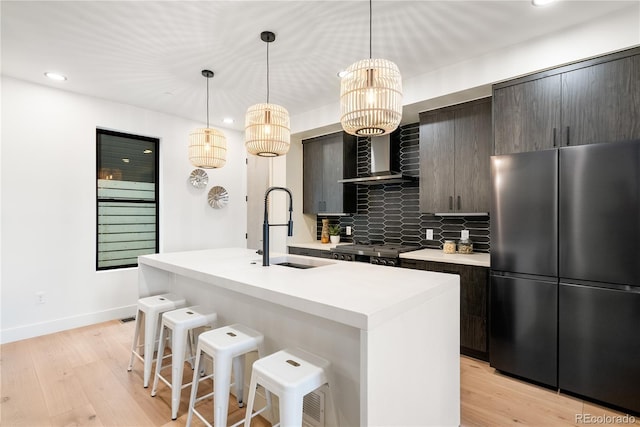 kitchen featuring sink, a center island with sink, stainless steel fridge, decorative backsplash, and wall chimney range hood