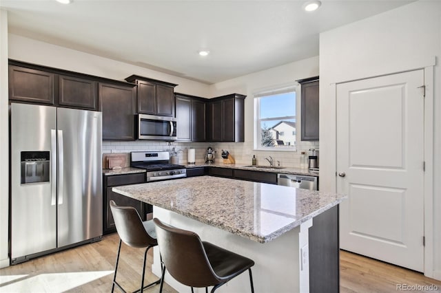 kitchen featuring dark brown cabinetry, a center island, light stone countertops, stainless steel appliances, and light wood-type flooring