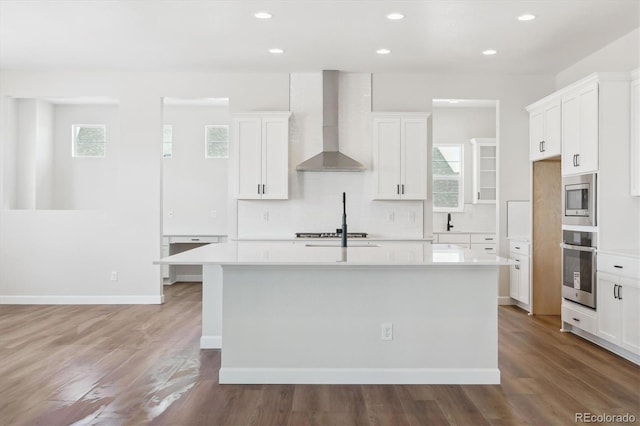 kitchen featuring stainless steel appliances, a kitchen island with sink, a healthy amount of sunlight, and wall chimney range hood