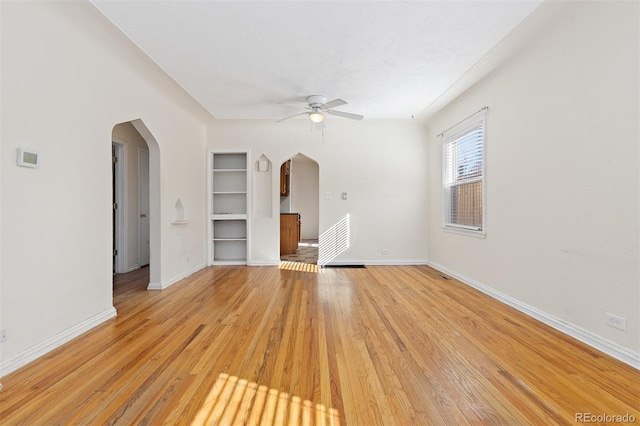 spare room with baseboards, arched walkways, built in features, ceiling fan, and light wood-type flooring