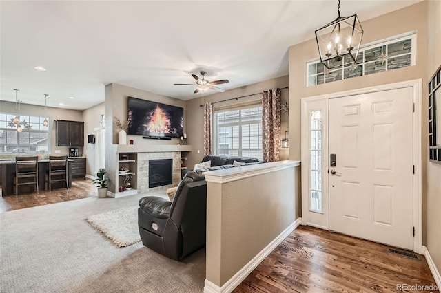 foyer entrance featuring ceiling fan with notable chandelier, wood finished floors, visible vents, baseboards, and a glass covered fireplace