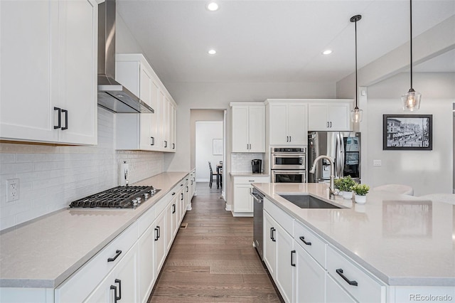 kitchen with wall chimney range hood, a kitchen island with sink, appliances with stainless steel finishes, a sink, and dark wood-style floors