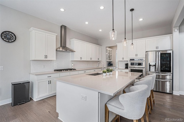 kitchen featuring wall chimney range hood, a sink, an island with sink, stainless steel appliances, and recessed lighting