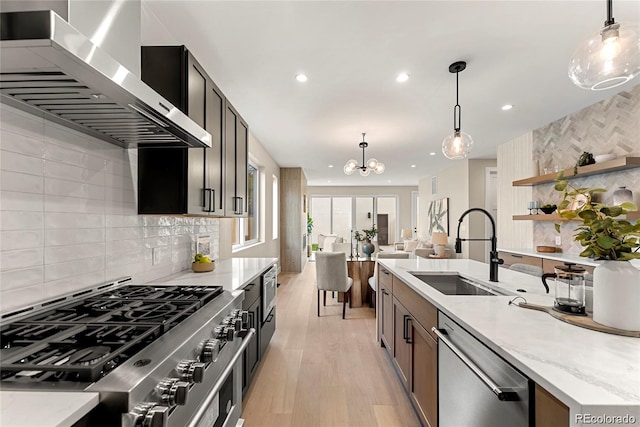 kitchen with light stone countertops, a sink, appliances with stainless steel finishes, pendant lighting, and wall chimney range hood