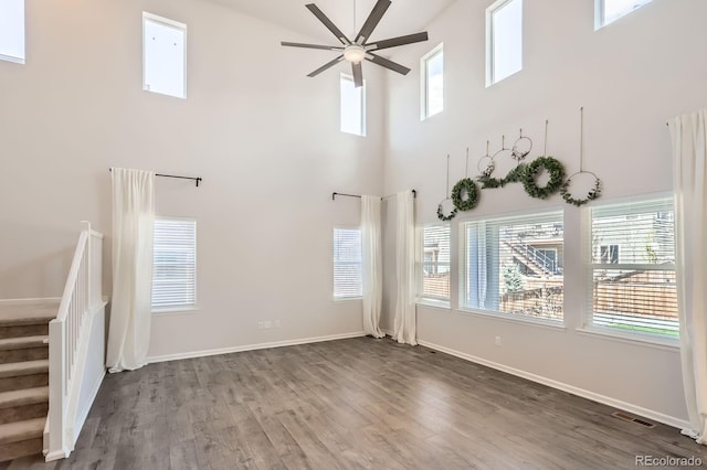 unfurnished living room featuring ceiling fan, wood finished floors, a towering ceiling, visible vents, and stairway