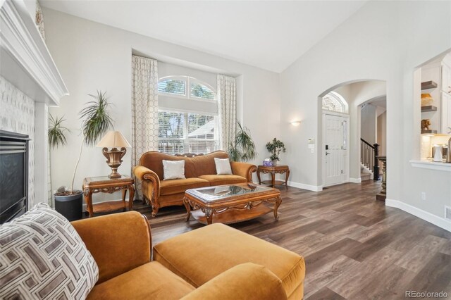 living area featuring arched walkways, high vaulted ceiling, a glass covered fireplace, baseboards, and wood finished floors