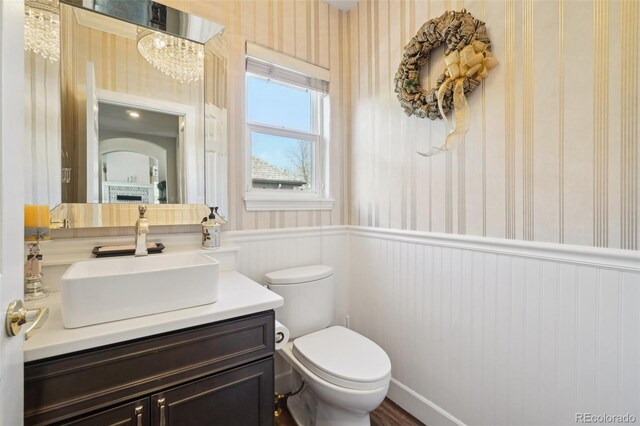 half bath featuring wainscoting, vanity, toilet, a chandelier, and wallpapered walls