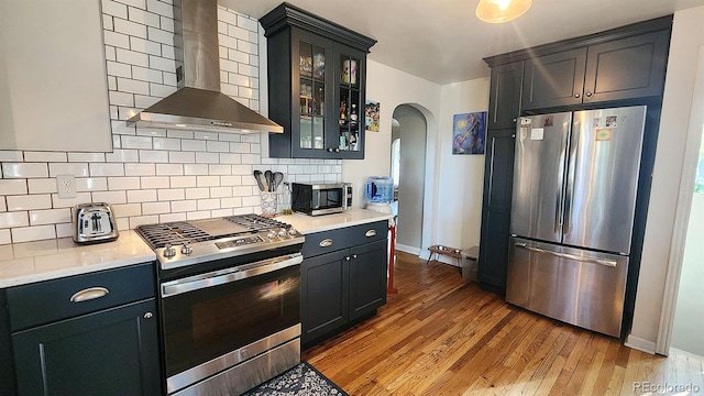 kitchen featuring decorative backsplash, appliances with stainless steel finishes, wall chimney range hood, and light hardwood / wood-style floors