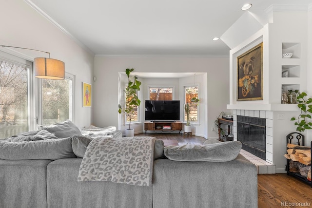 living area featuring crown molding, built in shelves, dark wood-type flooring, and a tiled fireplace