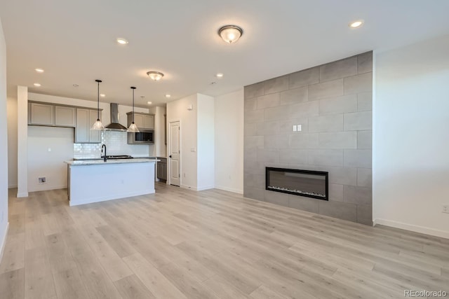 kitchen with gray cabinetry, a tile fireplace, wall chimney range hood, an island with sink, and light hardwood / wood-style floors