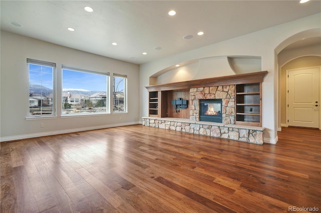 unfurnished living room with arched walkways, a stone fireplace, recessed lighting, wood finished floors, and baseboards