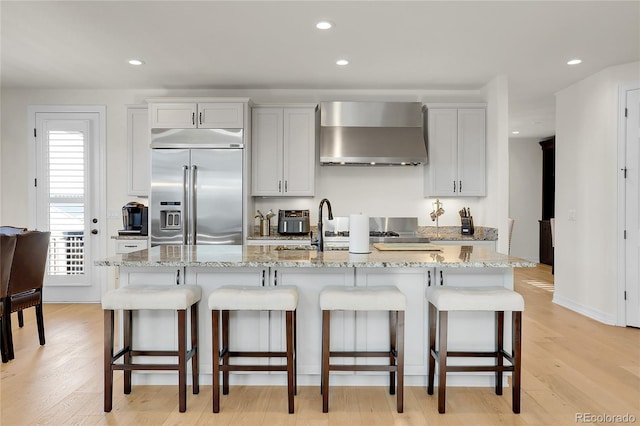 kitchen featuring stainless steel built in refrigerator, a breakfast bar, white cabinetry, an island with sink, and wall chimney range hood