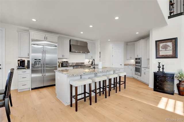 kitchen featuring wall chimney exhaust hood, appliances with stainless steel finishes, a kitchen island with sink, and white cabinets