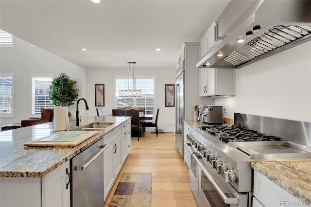 kitchen with appliances with stainless steel finishes, sink, wall chimney range hood, light stone counters, and a center island with sink