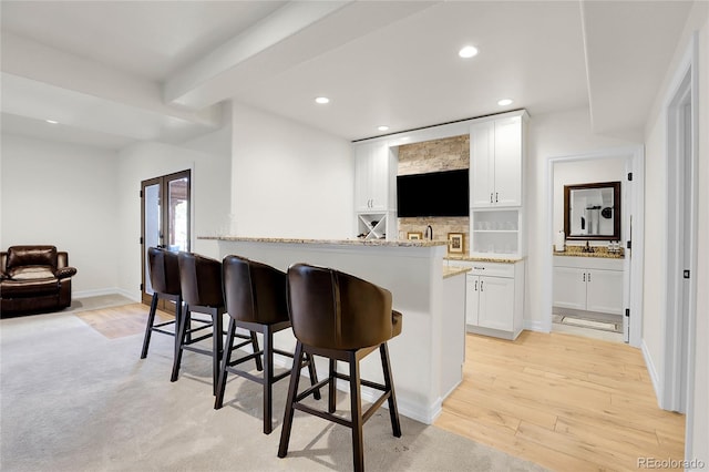 kitchen with light stone countertops, light hardwood / wood-style floors, white cabinets, a kitchen bar, and beamed ceiling