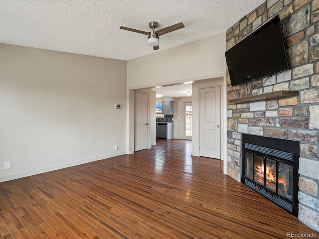 unfurnished living room with a textured ceiling, a stone fireplace, a ceiling fan, baseboards, and wood finished floors