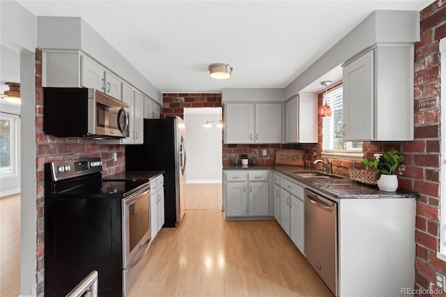 kitchen with light wood finished floors, brick wall, appliances with stainless steel finishes, dark stone countertops, and a sink