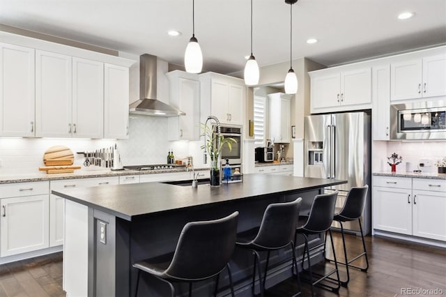 kitchen featuring appliances with stainless steel finishes, wall chimney exhaust hood, a kitchen island with sink, white cabinets, and hanging light fixtures