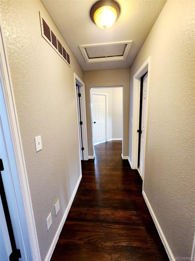 hall featuring a textured wall, visible vents, baseboards, and wood finished floors