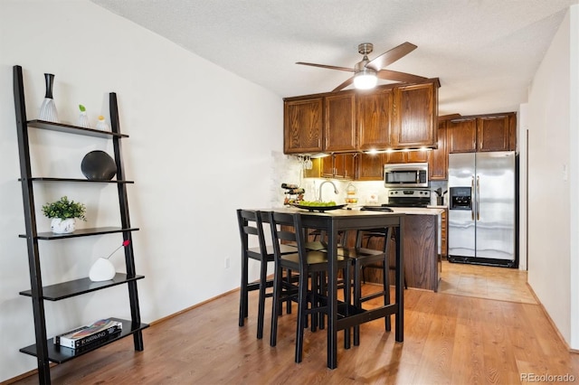 kitchen with light wood-type flooring, ceiling fan, kitchen peninsula, appliances with stainless steel finishes, and a kitchen bar