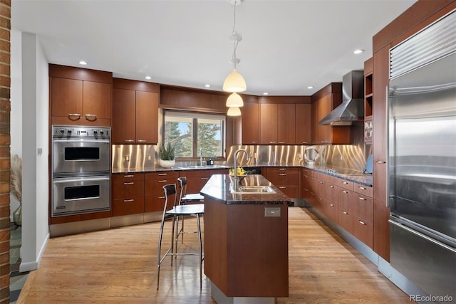 kitchen with an island with sink, modern cabinets, appliances with stainless steel finishes, wall chimney range hood, and a sink