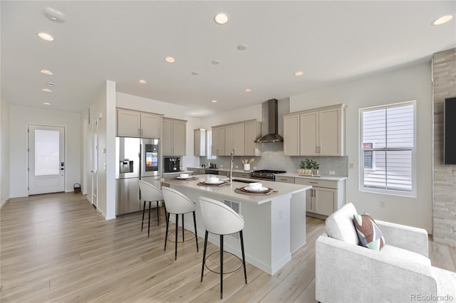 kitchen with wall chimney exhaust hood, light wood-type flooring, a breakfast bar, an island with sink, and stainless steel appliances