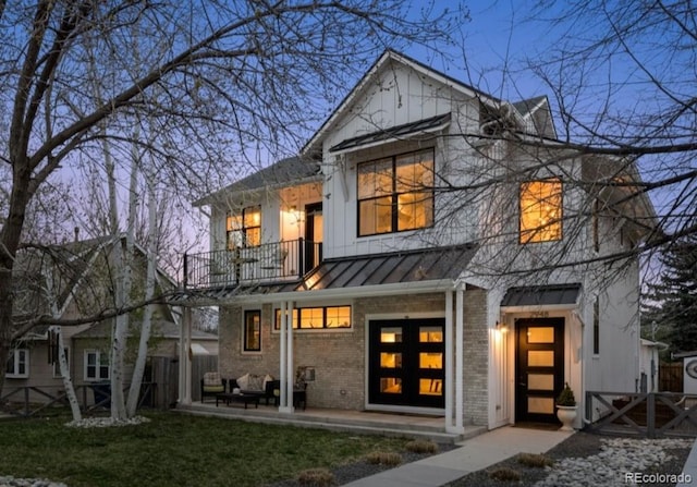 back of house at dusk with fence, brick siding, a balcony, a standing seam roof, and a yard