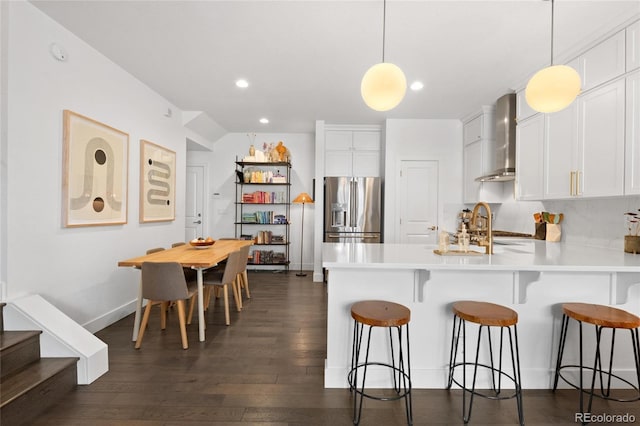 kitchen featuring wall chimney range hood, white cabinets, pendant lighting, and stainless steel refrigerator with ice dispenser