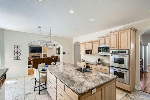 7482 Routt Ln, Arvada, CO 80005 - kitchen featuring stainless steel appliances, arched walkways, light brown cabinets, a center island with sink, and light stone countertops