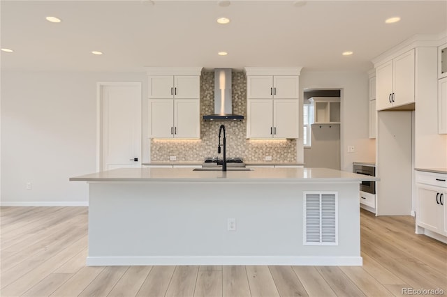 kitchen with white cabinets, a kitchen island with sink, wall chimney exhaust hood, and light wood-type flooring