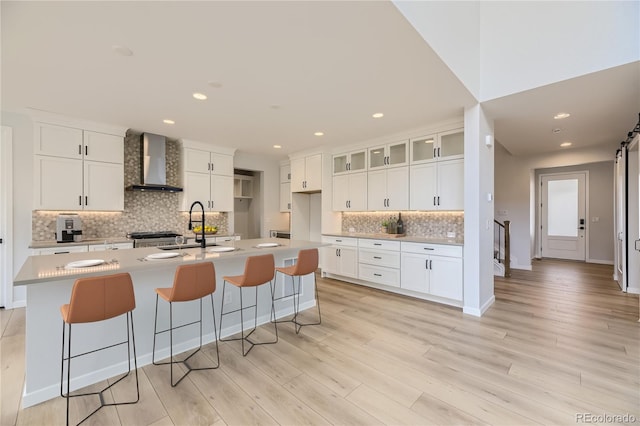 kitchen with wall chimney range hood, sink, an island with sink, white cabinetry, and a breakfast bar area