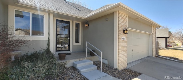 view of exterior entry with stucco siding, concrete driveway, an attached garage, a shingled roof, and brick siding