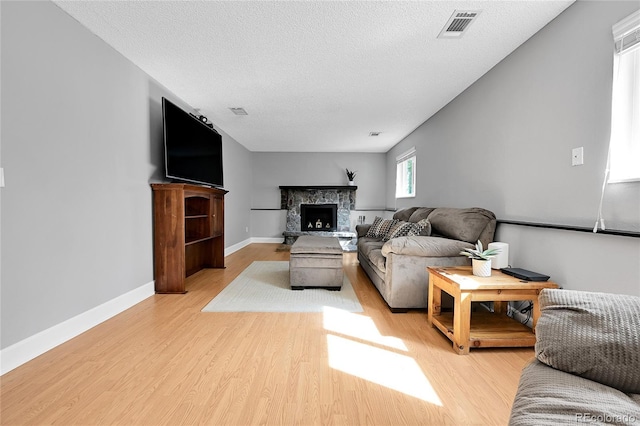 living room featuring light wood-type flooring, visible vents, a textured ceiling, a stone fireplace, and baseboards