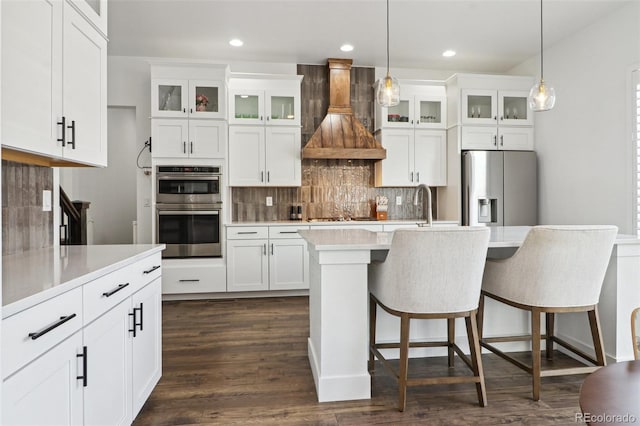 kitchen with an island with sink, dark wood finished floors, appliances with stainless steel finishes, decorative backsplash, and custom range hood