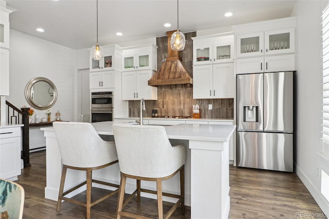 kitchen with appliances with stainless steel finishes, premium range hood, decorative backsplash, light countertops, and an island with sink