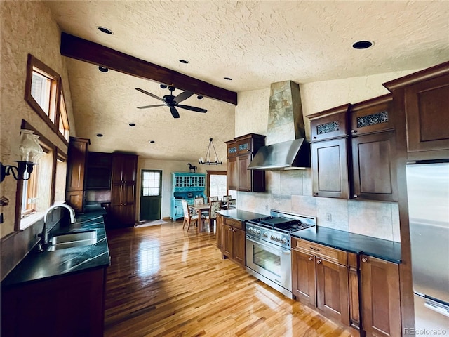 kitchen featuring beam ceiling, stainless steel appliances, dark countertops, a sink, and wall chimney exhaust hood