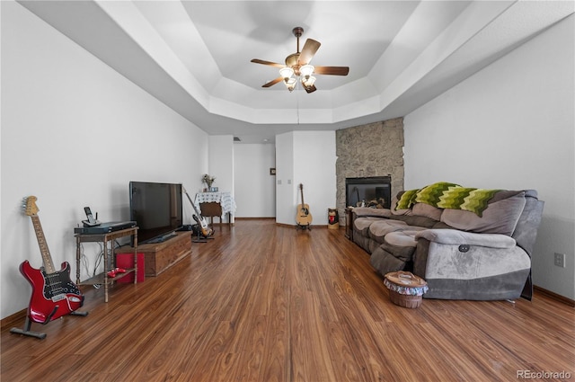 living room featuring a stone fireplace, wood-type flooring, a raised ceiling, and ceiling fan