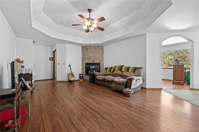 living room featuring a stone fireplace, ceiling fan, dark wood-type flooring, and a raised ceiling