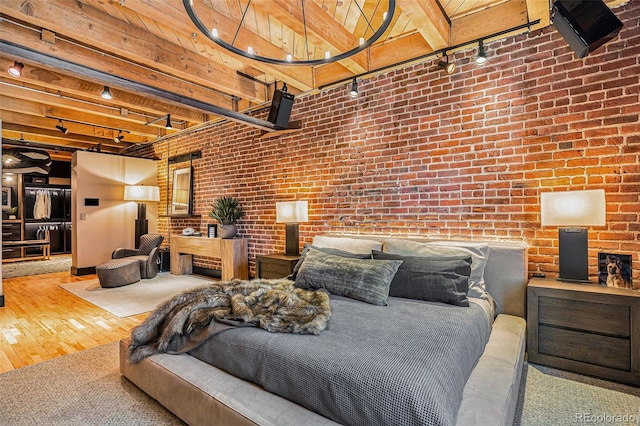 bedroom with track lighting, beamed ceiling, a towering ceiling, brick wall, and hardwood / wood-style floors