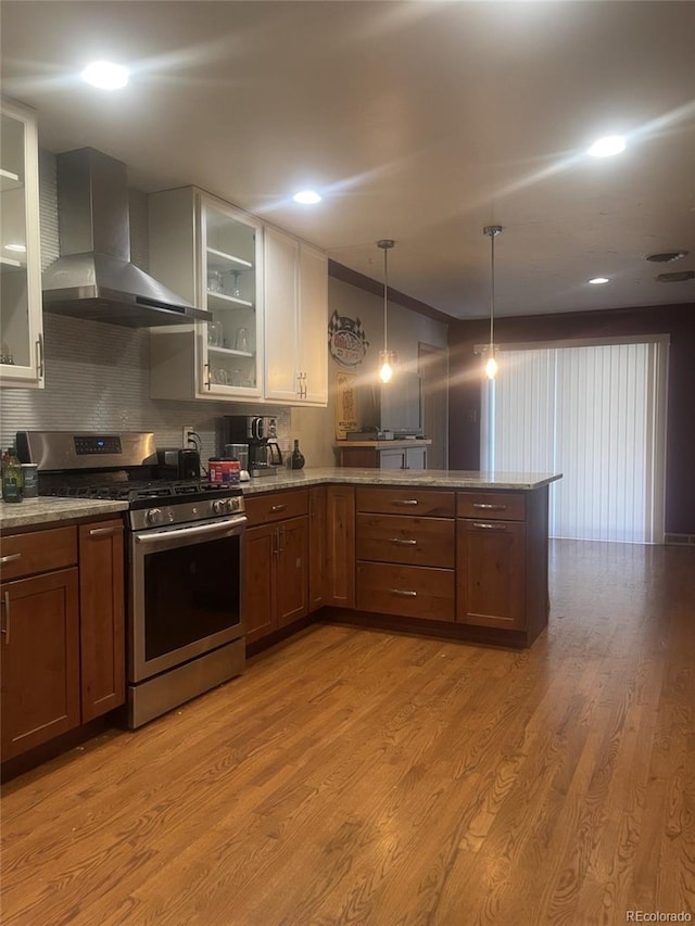 kitchen with stainless steel gas stove, hanging light fixtures, wall chimney range hood, backsplash, and light wood-type flooring