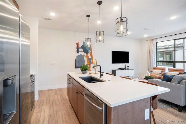 kitchen with light wood-type flooring, a center island with sink, appliances with stainless steel finishes, decorative light fixtures, and sink