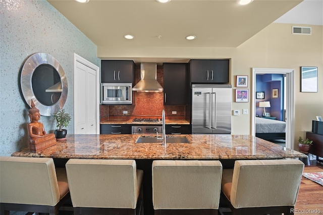 kitchen featuring visible vents, light stone counters, built in appliances, wall chimney range hood, and a sink