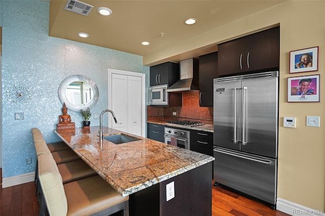 kitchen with wallpapered walls, visible vents, wall chimney exhaust hood, built in appliances, and a sink