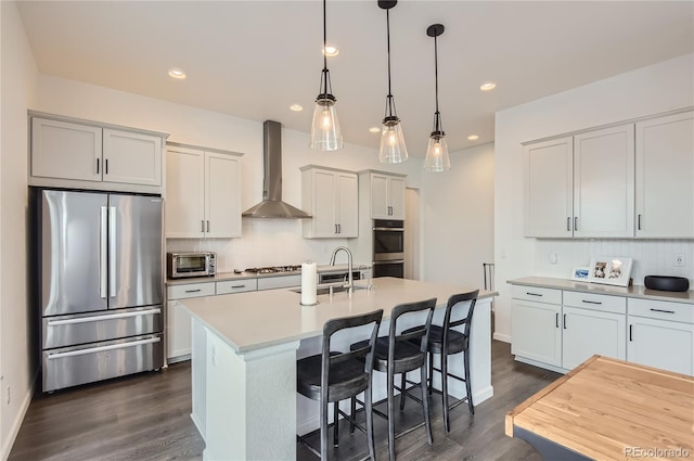 kitchen with appliances with stainless steel finishes, hanging light fixtures, dark wood-type flooring, wall chimney exhaust hood, and a center island with sink