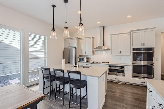 kitchen featuring an island with sink, stainless steel appliances, decorative light fixtures, dark hardwood / wood-style floors, and wall chimney range hood