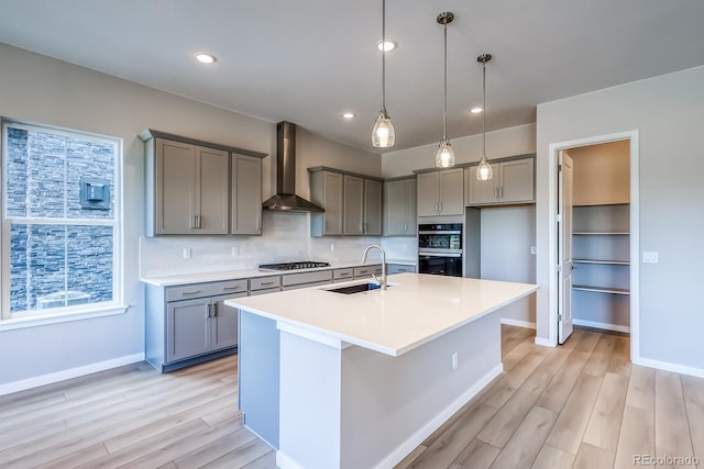 kitchen featuring wall chimney exhaust hood, stainless steel gas cooktop, sink, light wood-type flooring, and a kitchen island with sink