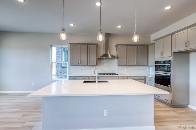 kitchen with hanging light fixtures, an island with sink, sink, and wall chimney range hood