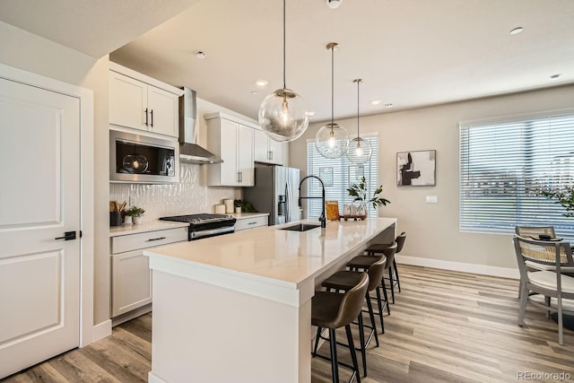 kitchen with white cabinetry, appliances with stainless steel finishes, wall chimney exhaust hood, and an island with sink