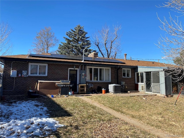 rear view of house with a chimney, a yard, central air condition unit, roof mounted solar panels, and brick siding